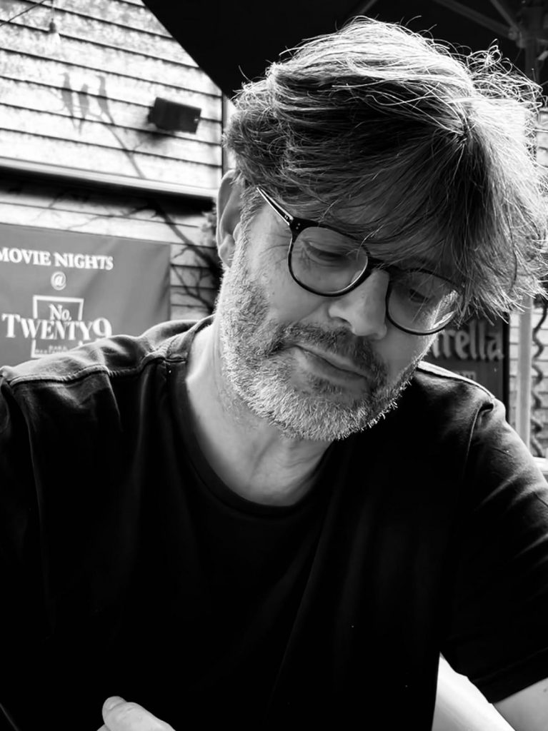 Black and white photograph of a middle-aged man, seated. He is wearing a black t-shirt and dark-rimmed glasses. He has floppy dark hair, slightly greying, and a stubbly beard. He is looking down, a half-smile across his face.